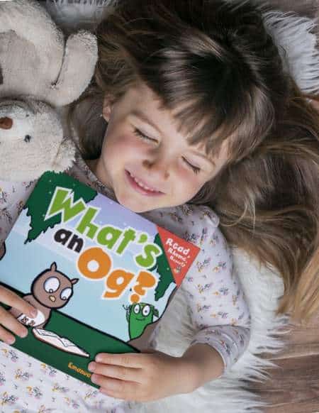 Young girl is lying down on a furry rug with her eyes closed holding a copy of "What's an Og?" close to her chest. She is smiling. She has long brown hair and a teddy bear beside her ear.