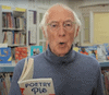 Shows Roger Mcgough a greyhaired man with transparent round reading glasses. He is reading a book "Poetry Pie" and is wearing a blue-grey Polo-neck sweater. He is standing in front of bookshelves and rows of books.